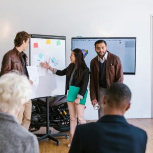 A multicultural team collaborates in an office, presenting ideas using a whiteboard and digital screen.
