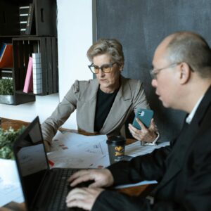 Two senior colleagues discussing a project in a modern office setting, using laptops and mobile devices.
